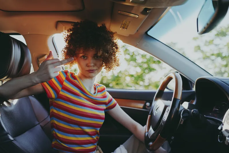 Cheerful woman inside car, striped colorful shirt, sunlit moment, relaxed driver posture, retro film color, natural light, trees outside, candid lifestyle scene