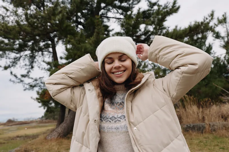 Smiling woman in a beige winter coat and knit sweater outdoors, posing in a windy countryside with trees and grassy field.