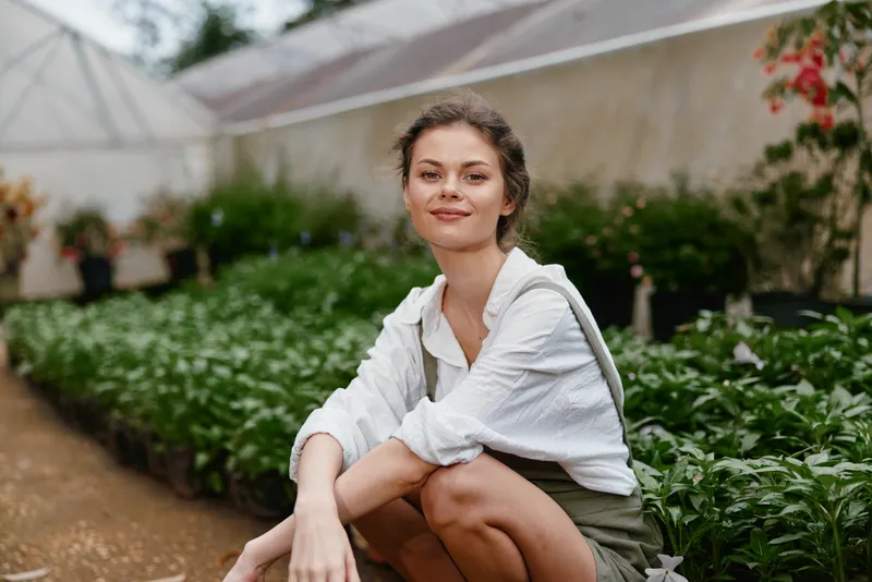 Young woman in a greenhouse, wearing a casual outfit, smiling amidst lush greenery, symbolizing a connection with nature and healthy living