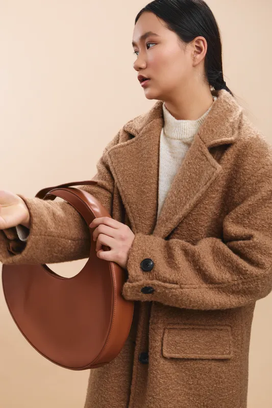 asian woman wearing a warm coat holds a round handbag in a neutral studio, exuding calm confidence and subtle sophistication, creating a stylish contemporary mood suitable for fashion and lifestyle
