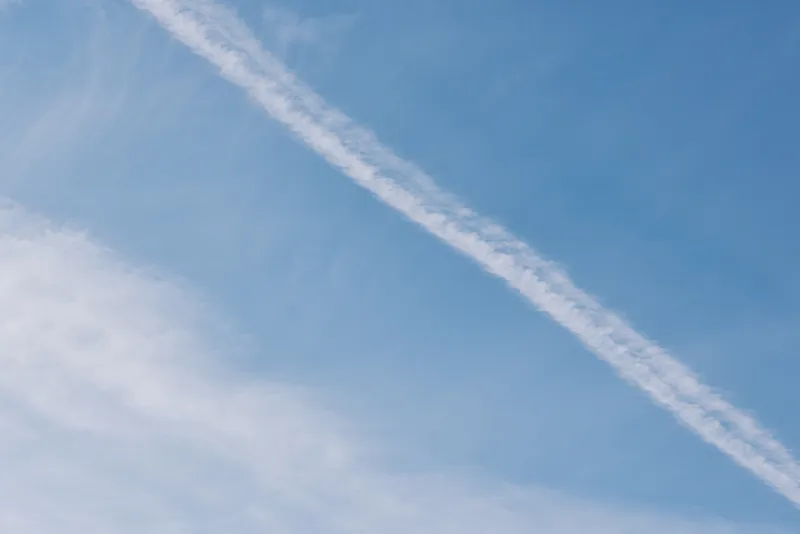 Blue sky with a white contrail streak through light clouds, creating a calm daytime scene ideal for travel, aviation, or nature backdrops.