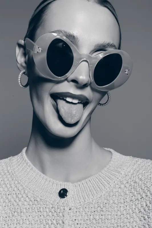 Happy white woman with light skin wearing large round sunglasses and hoop earrings sticks out tongue. Close-up portrait in knitted sweater on plain background.