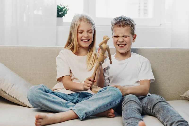 Two laughing children sit on a couch playing with a wooden figure, enjoying a playful moment in a bright living room with casual clothes and natural light.