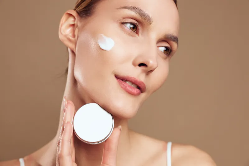 Young woman applying face cream on cheek holding jar close to skin, natural skincare routine, smooth complexion, soft lighting, neutral background, beauty care concept