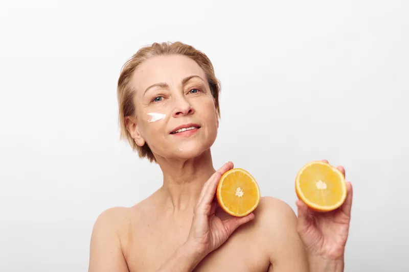 Mature woman with natural skin holds two orange halves near face with cream on cheek. Concept of healthy skincare, natural beauty, vitamin C, and organic self-care in bright studio light.