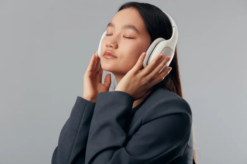 Young woman wearing white headphones, eyes closed and enjoying music with relaxed expression, casual dark jacket on grey background, portrait of peaceful listening moment.