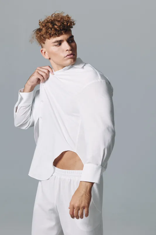 Young man with curly hair wearing white shirt and pants posing in studio, fashion portrait with casual style and neutral background.