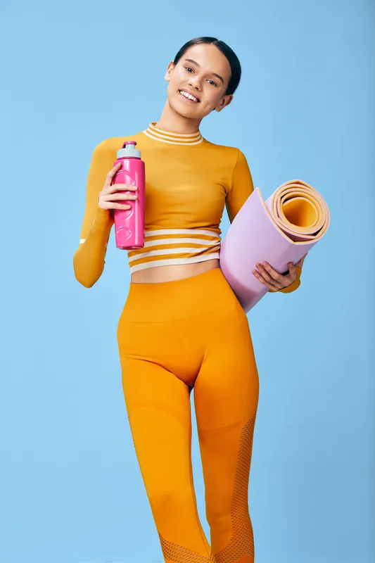 Woman fitness enthusiast in bright orange activewear, holding a pink water bottle and a rolled yoga mat, set against a blue studio backdrop, ready for a workout and health routine.