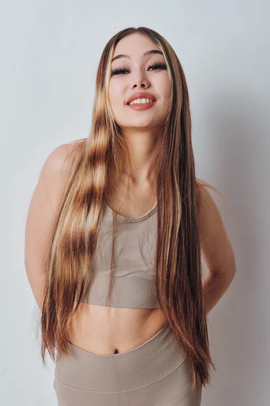 Young woman with long hair and smiling face wearing sportswear standing with hands behind back against plain background. Healthy lifestyle concept with confident posture.