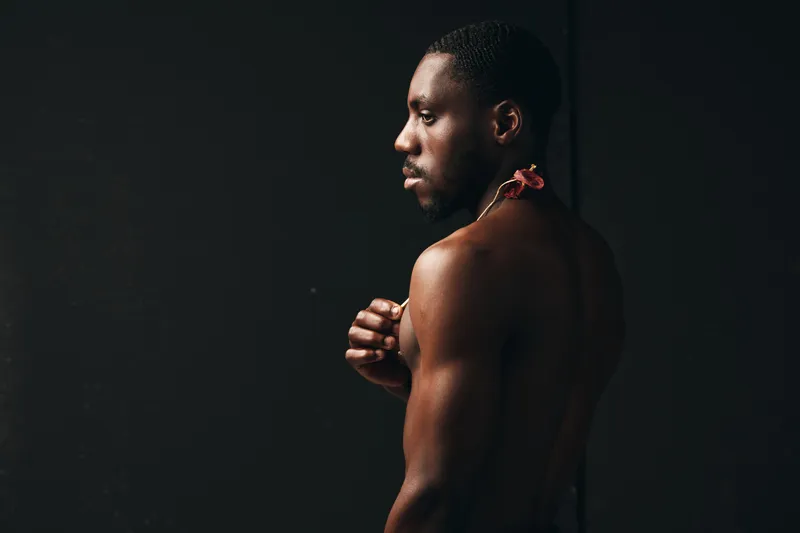 Creative visual designer style portrait of African American man with dark skin in profile, bare torso and flower on shoulder against black background in studio lighting.