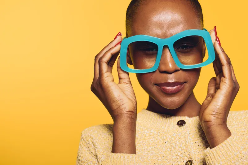 Close-up of a smiling young black woman with dark skin wearing oversized blue glasses and a beige sweater against a yellow background. Concept of fashion and joyful expression.
