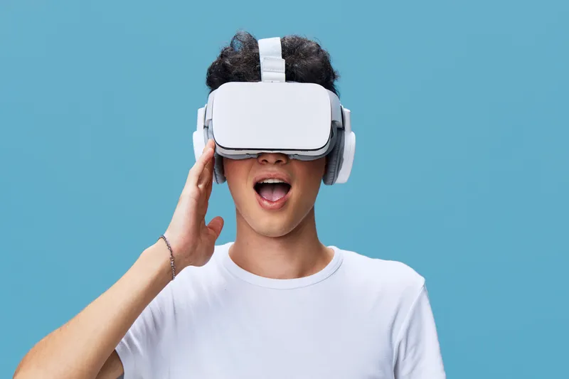 young man wearing white virtual reality headset and headphones against blue background, experiencing immersive technology, casual clothing, entertainment concept