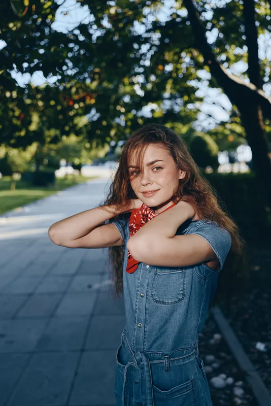 young woman smiling outdoors wearing denim dress and red scarf under green trees on sunny day with natural light and urban background