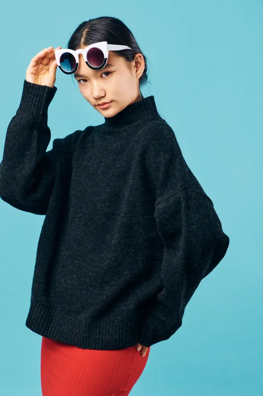 Young woman in black oversized sweater and red skirt adjusting white sunglasses on light blue background. Fashion pose with confident expression and casual trendy outfit studio portrait.