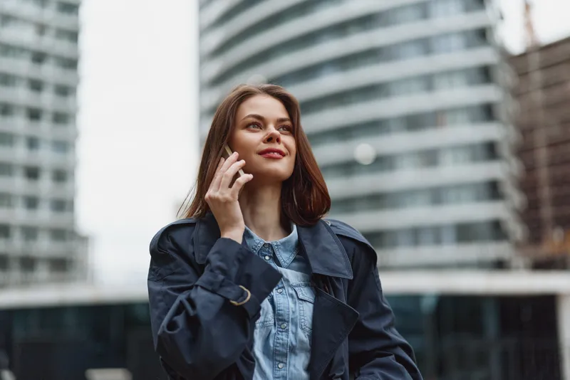 A young woman on a phone speaks in a cityscape, wearing a denim jacket over a denim shirt, standing beside modern glass buildings in daylight, captured in a natural candid moment.