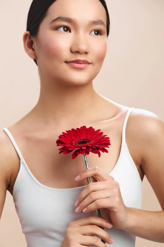 asian woman holds a vivid red flower in a clean studio setting, smiling softly as she gazes slightly away, projecting calm elegance, warmth, and a graceful, contemporary mood.