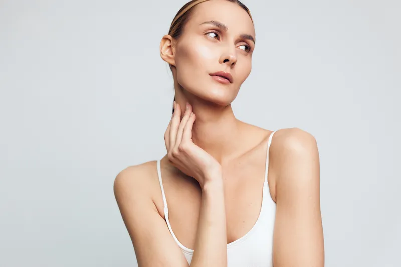 Portrait of a young woman with clear skin and slicked back hair wearing a white tank top, posing with her hand on her neck against a plain light background. Beauty, skin care.