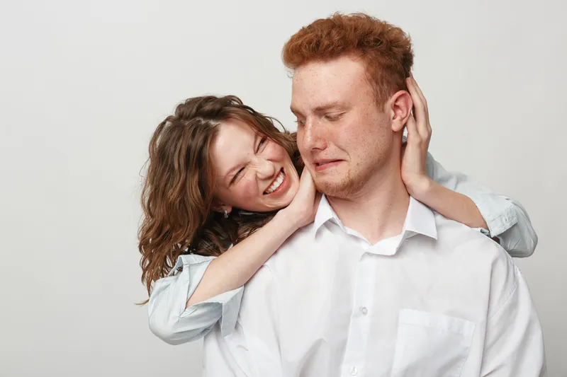 Playful young woman with curly hair hugs smiling man with red hair from behind. Both wear casual white shirts, expressing joy and affection in a bright studio setting. Concept of love and friendship.
