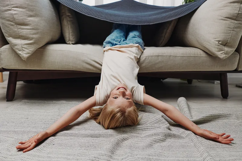 Happy child lying upside down on carpet with arms spread, smiling under sofa blanket in cozy living room. Concept of playful childhood, comfort, and home leisure activity.