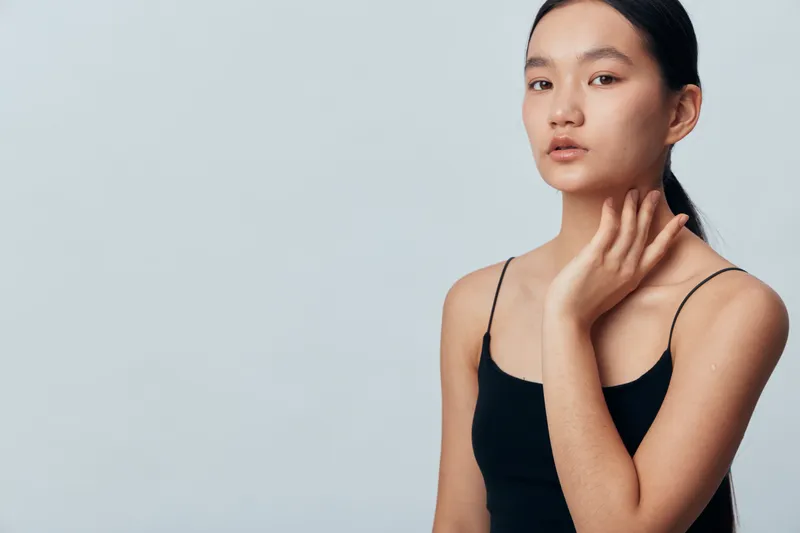 Young woman with clear skin and black hair wears black sleeveless top posing with hand near neck against light gray background for beauty and skincare concept.