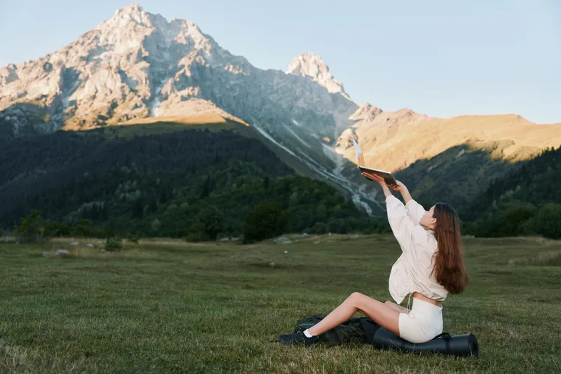 A woman sits on a grassy meadow beneath towering mountains, launching a kite. She smiles warmly, enjoying fresh air, wide horizon, and a serene outdoor atmosphere in the scenic landscape.