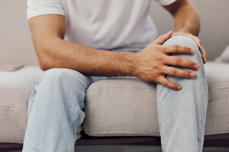 Young man sitting on a sofa, displaying a thoughtful expression while holding his knee. Warm tones and cozy decor create a relaxed atmosphere.