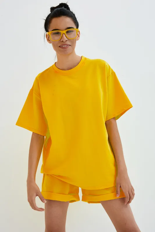 Young woman wearing bright yellow casual outfit and matching glasses stands confidently against a white background, showcasing cheerful summer style and relaxed mood.