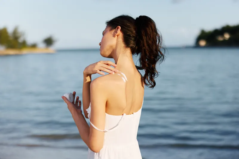 A serene portrait of a woman standing by the sea in a white sundress, sunlight on bare shoulders, gentle breeze, and tranquil water in the background.