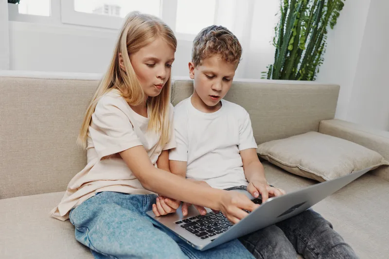 Curious children sitting on a sofa, focused on a laptop screen together. Casual home setting with natural light, illustrating learning, technology, and sibling interaction concepts.