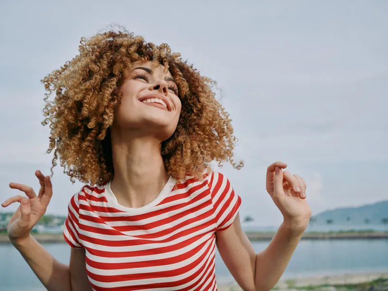 A joyful woman with curly hair smiles toward the sky, wearing a red striped t-shirt, standing by a calm shore with distant hills and soft blue sky.