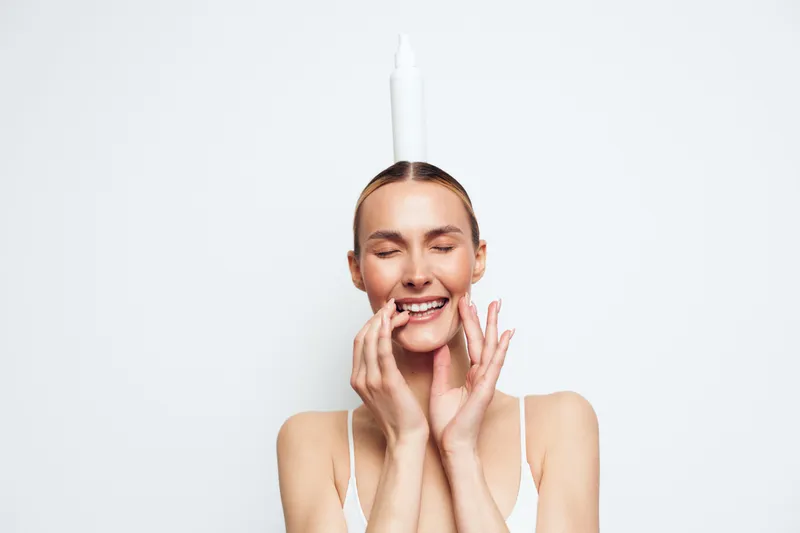 Young woman with closed eyes smiling gently, balancing white bottle on head, minimalistic background, natural light, relaxed and playful mood, skincare concept