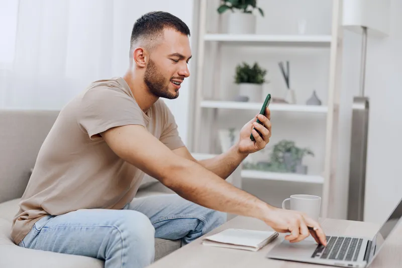 Smiling man engaging with smartphone while working on laptop, showcasing a modern lifestyle and digital connectivity, perfect for remote work themes in 2025.