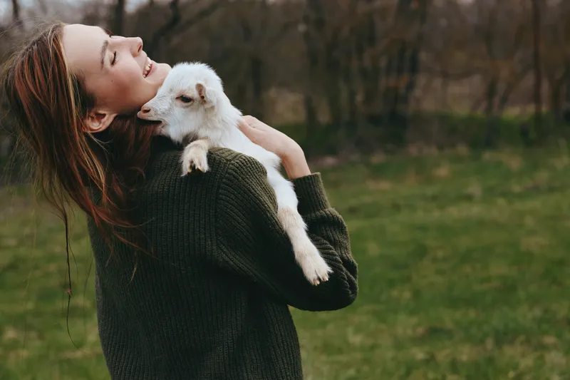 Cheerful woman outdoors, holding a goat kid close, sunlit moment, relaxed posture, soft breeze, natural colors, lush grass background, candid, moment, everyday, life, outdoor, scenery, rustic, vibe