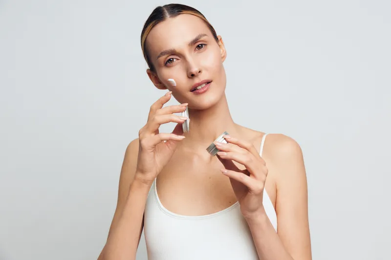 Woman skincare serum face beauty bottle hygiene spa studio moisturizer, female applies serum to her clean face in studio portrait, minimal light gray background, holds bottle and spray, natural glow,