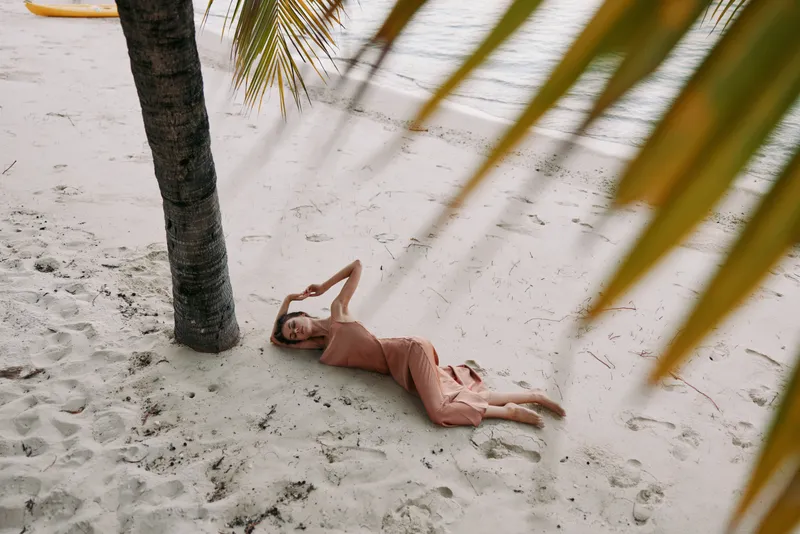 A lone person lies on soft beach sand under a palm tree, sunlit calm, relaxed pose, conveying serenity, warmth, and a tranquil tropical escape.