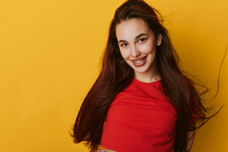 Young woman with long hair smiling and posing in red shirt on yellow background. Portrait of happy female model with natural makeup and casual style in studio lighting.