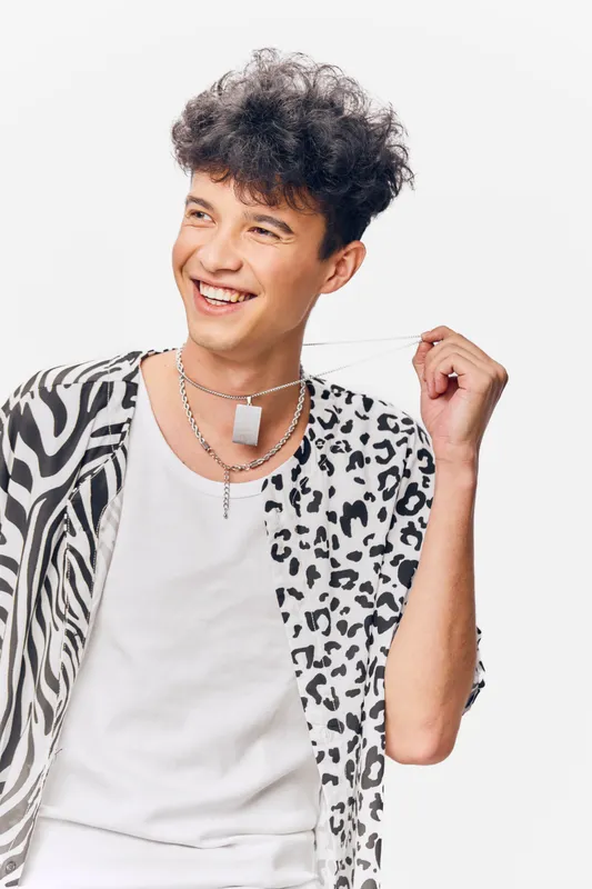 smiling man wearing patterned shirt and white tank top, showing playful attitude with curly hair and casual silver necklaces against white background