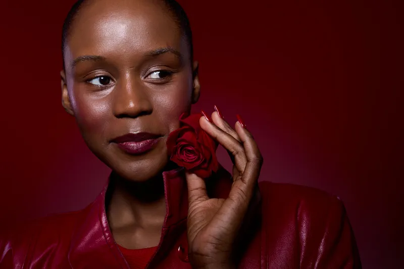 Close-up of a dark skinned African woman with short hair holding a red rose near her face. She wears a red leather jacket against a red background. Concept of beauty, elegance, and confidence.