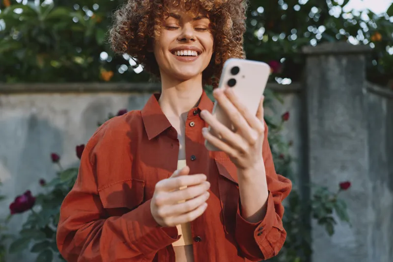 Bright outdoor lifestyle scene with a smiling woman holding a smartphone, casual fashion, garden backdrop, natural light, candid moment, relaxed vibe, colorful surroundings