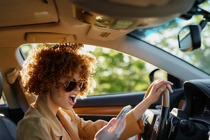 Young woman drives a car with curly hair and sunglasses laughs inside a sunlit warm interior, daylight filtering through the windshield creating a candid retro vibe