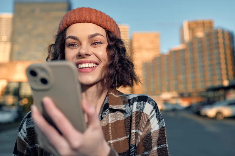 Woman smiling with smartphone on urban street at golden hour glow, candid lifestyle portrait emphasizing authenticity, mindful living and emotional storytelling in a modern city moment.
