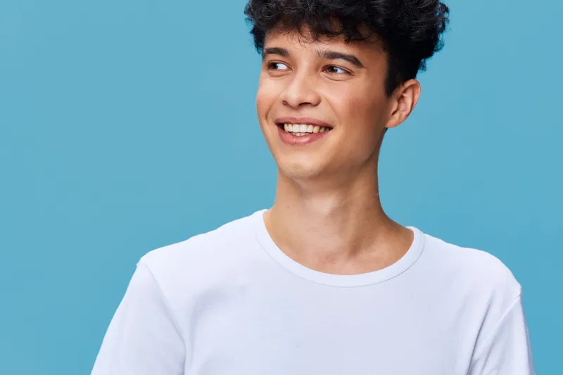 smiling young man wearing a white t-shirt looking aside against blue background, casual style, positive expression, fresh and bright lighting