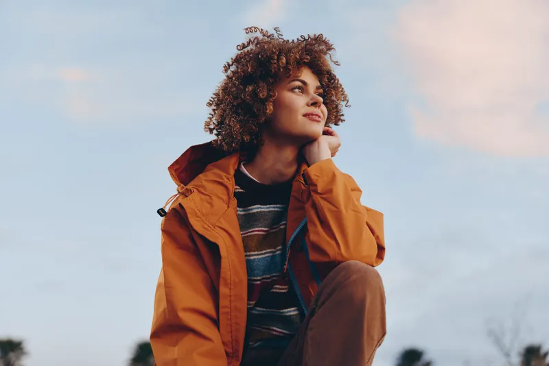 Woman outdoors lifestyle, curly hair, orange jacket, sunset light, thoughtful chin pose, warm breeze, casual fashion, relaxed expression, blurred sky and distant trees