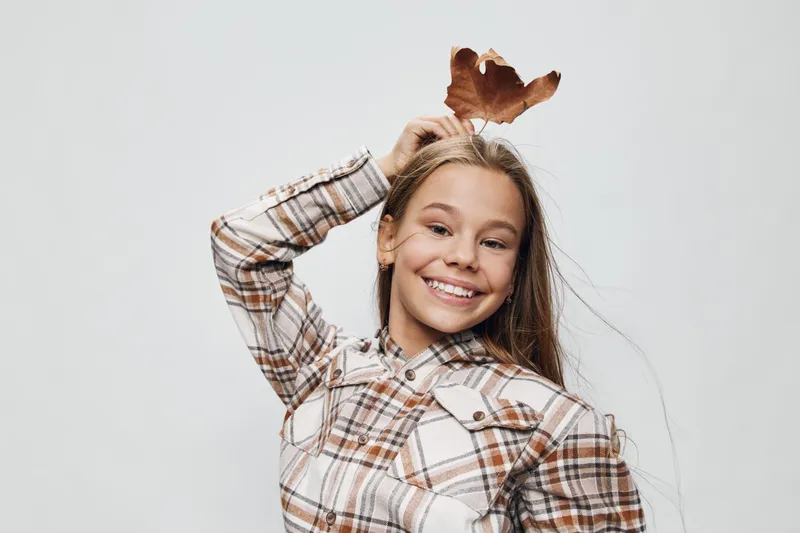 Girl smiling and holding a dry autumn leaf above her head wearing plaid shirt. Portrait of happy child with long hair and casual clothing in studio environment.