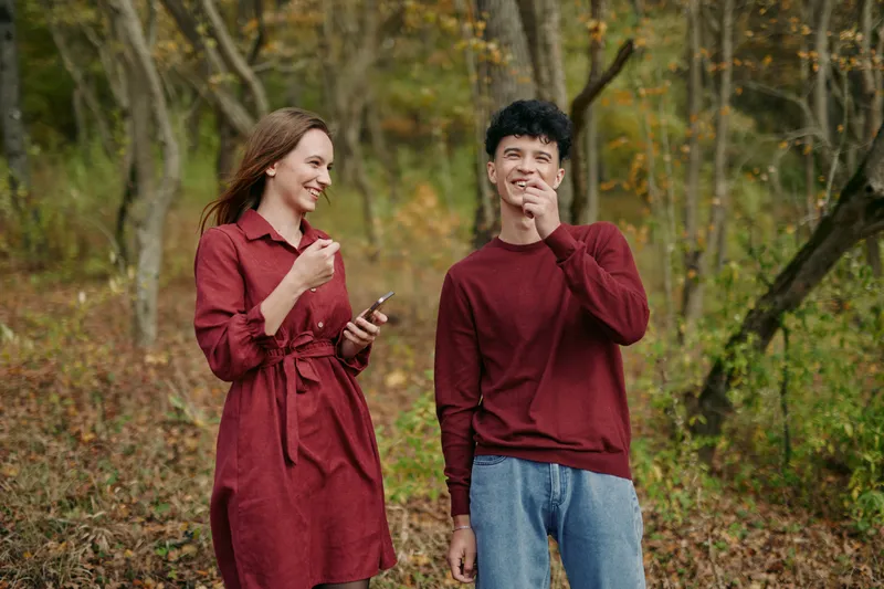 A candid scene in a autumn forest shows two friends in casual attire, smiling as one checks a phone. Subtle authenticity and credible emotion highlight a natural, unposed moment.