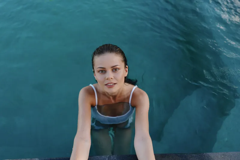 Young woman in a swimsuit climbing out of a swimming pool, wet hair, focused expression, clear blue water, summer lifestyle, outdoor activity, healthy living
