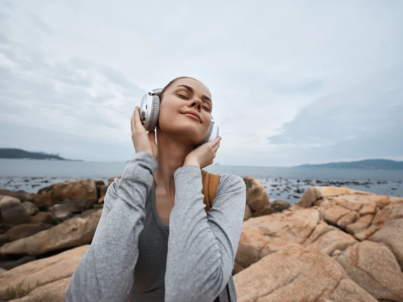 Young woman enjoying music on the rocky beach, wearing headphones and a casual outfit, with a serene expression and a cloudy sky background