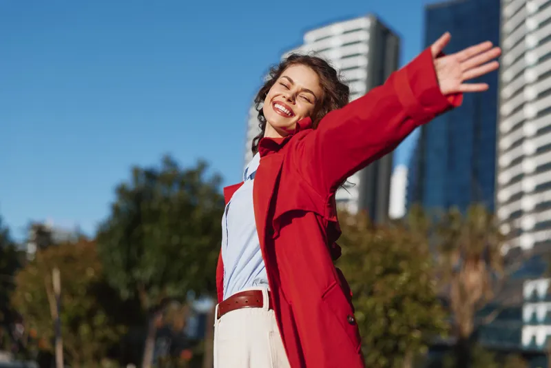 Cheerful woman outdoors, smiling with open arms, red coat fluttering, sunlit city scene, relaxed pose, vibrant colors, fresh breeze, carefree mood