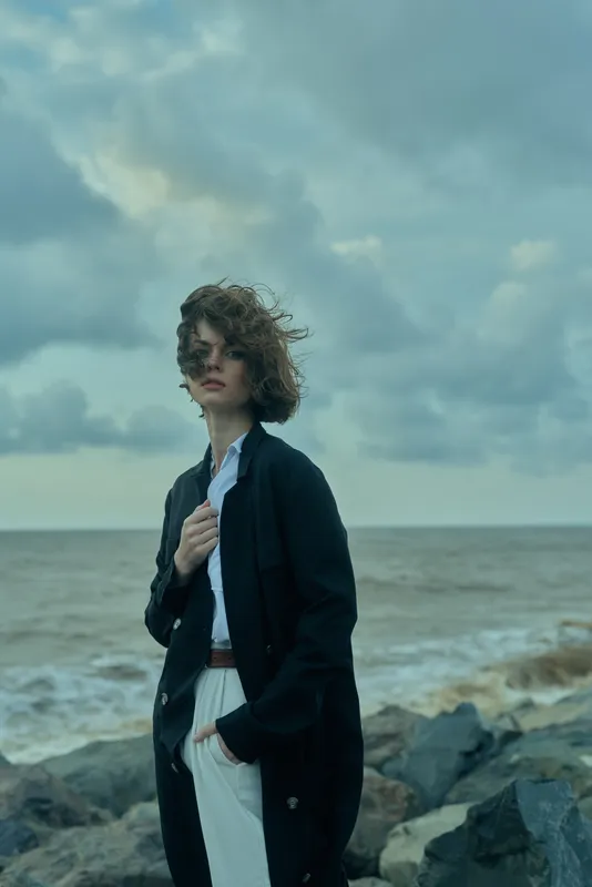 Seaside fashion on a windy coast with a stylish coat and relaxed shirt, dramatic ocean backdrop and rocky shoreline, a moody outdoor look for fashion storytelling