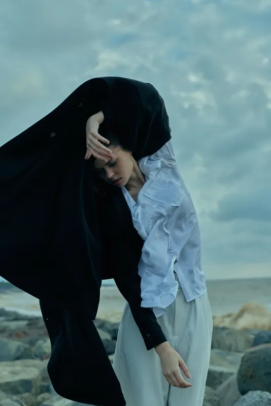 Veiled figure by the sea in a flowing black cloak and pale blue shirt, wind lifting fabric, tranquil coastline mood, mysterious elegance and motion at the shore